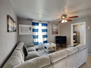 A living room with a grey sofa, a small white table, a TV, and a ceiling fan. - Surfside Escape by Ryson Vacation Rentals (Freeport)