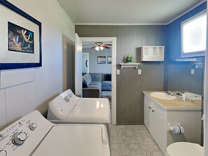 A laundry room with two washing machines, a small shelf with a decorative item, a bathroom with a sink and toilet, and a hallway leading to a living area. - Surfside Escape by Ryson Vacation Rentals (Freeport)