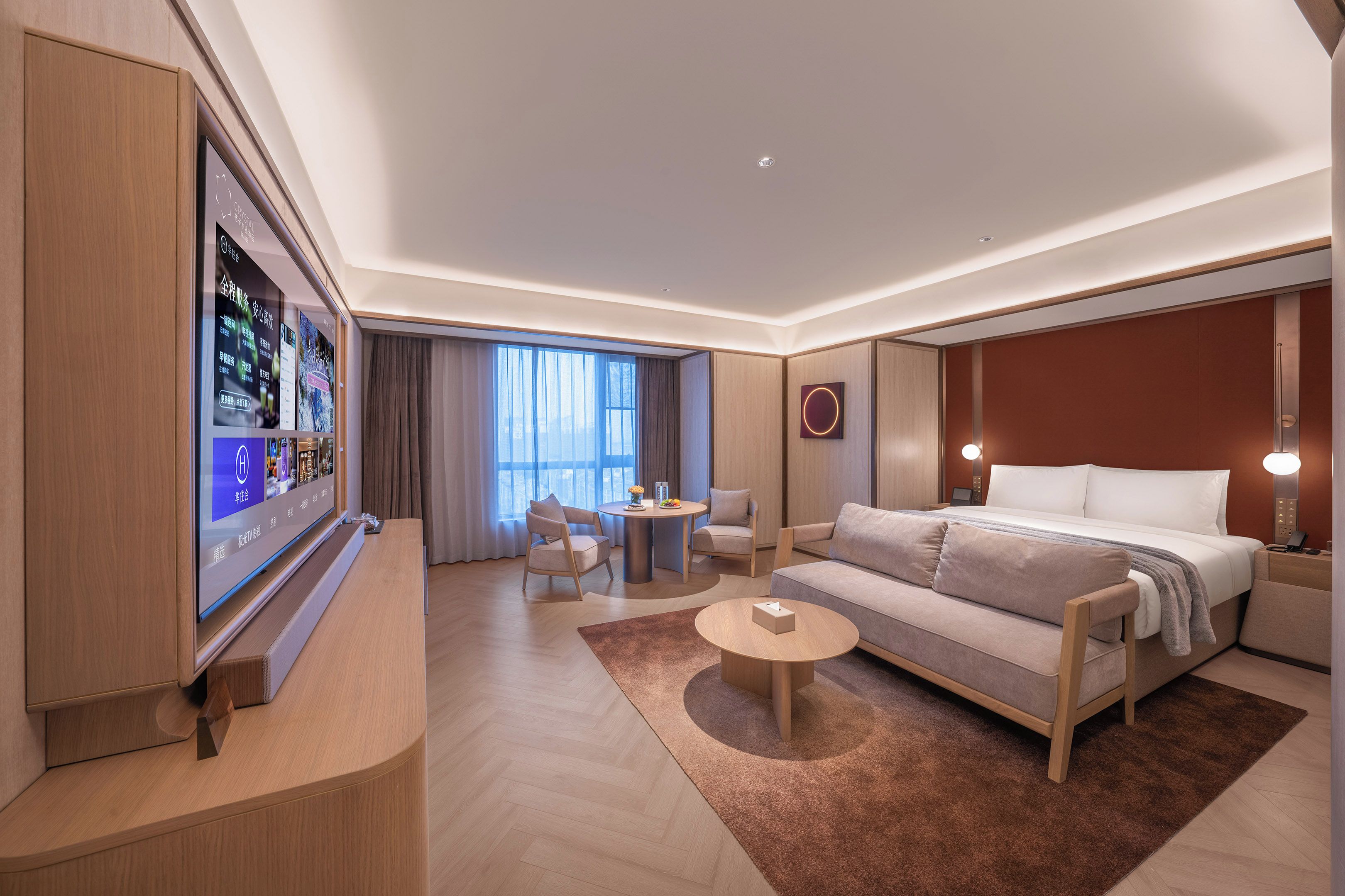 A modern hotel room with a large flat-screen TV, a sofa, a bed, a round coffee table, and a small side table.