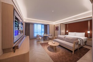 A modern hotel room with a large flat-screen TV, a sofa, a bed, a round coffee table, and a small side table.