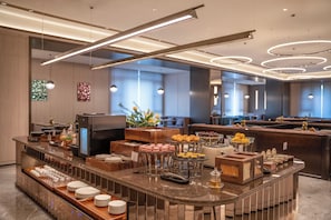 Un mostrador de desayuno moderno en un hotel, con diversas opciones de comida, una máquina de café y una zona de estar con vistas a la ciudad.