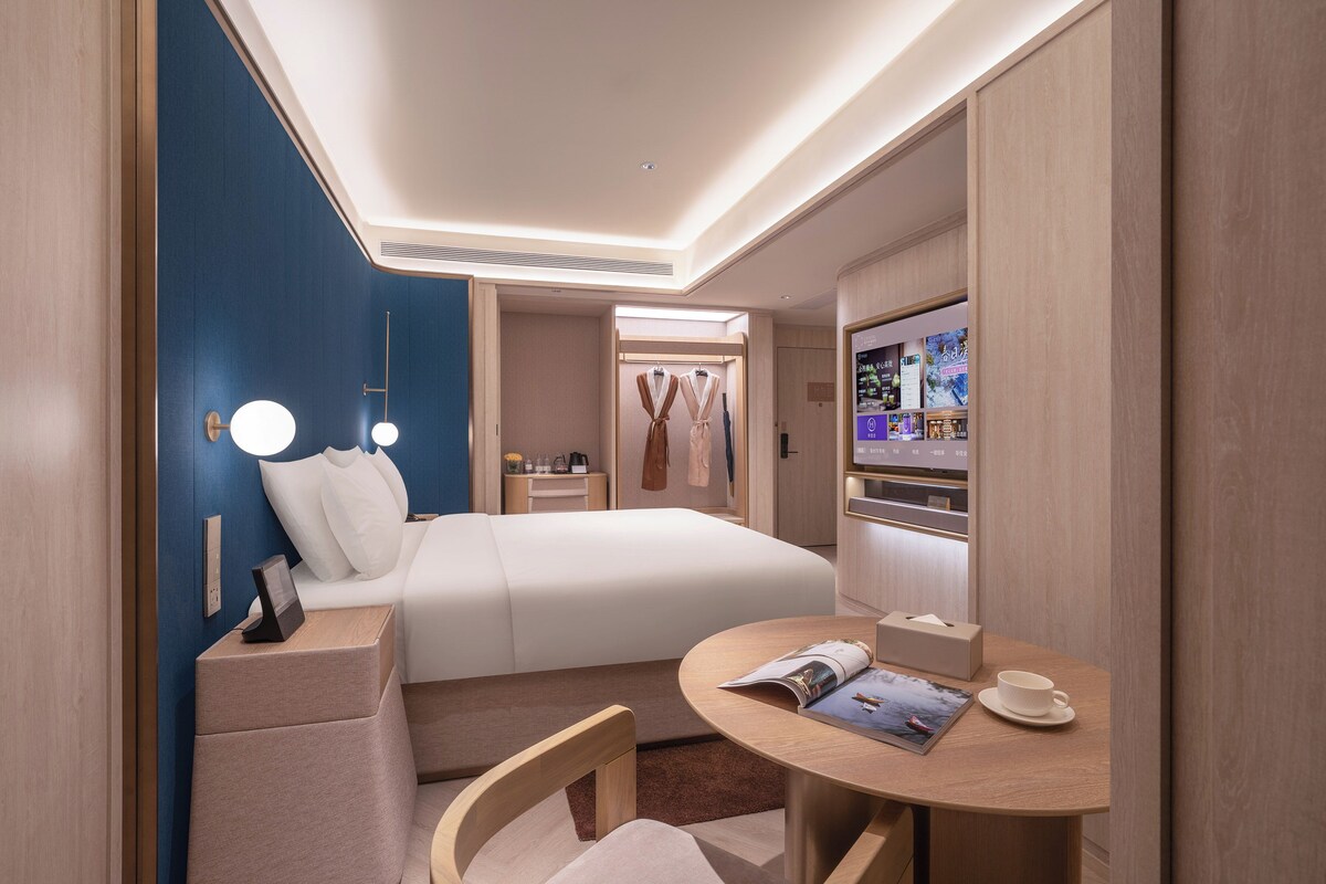Una habitación de hotel moderna con una cama, una mesita de noche, una lámpara y una pequeña mesa redonda con una taza y una revista.