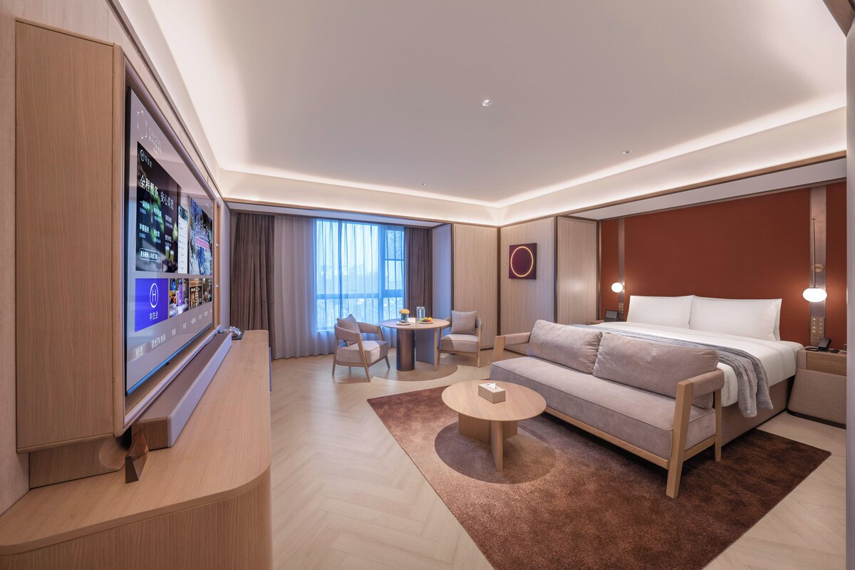 Una habitación de hotel moderna con un televisor de pantalla plana grande, un sofá, una cama, una mesa de centro y una pequeña mesa redonda con sillas.