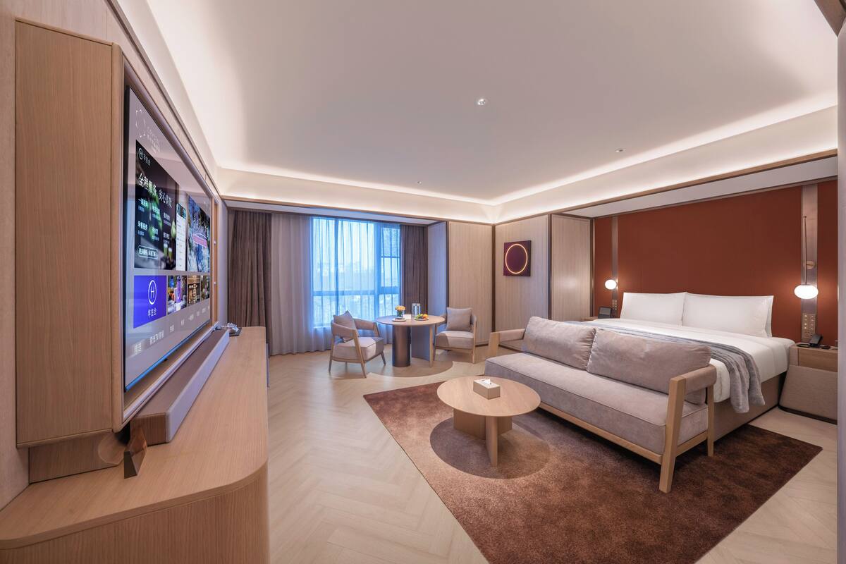 A modern hotel room with a large flat-screen TV, a sofa, a bed, a coffee table, and a small round table with chairs.