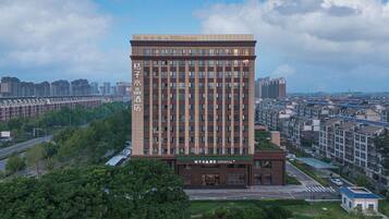 A modern hotel with a prominent name on the facade, surrounded by greenery and other buildings.