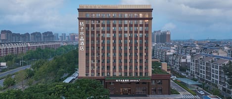 A modern hotel with a prominent name on the facade, surrounded by greenery and other buildings.