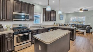 A modern kitchen with dark cabinets, stainless steel appliances, a central island, pendant lighting, and a view to the living area. - Bayview Oasis in the Heart of Seaside Heights (Seaside Heights)