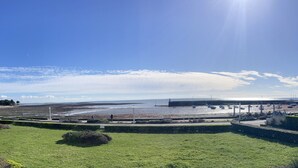 Un paysage côtier avec un ciel dégagé, un premier plan herbeux et une vue sur la mer, avec des bateaux et un phare visibles au loin.