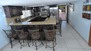 A kitchen with a central island, bar stools, dark cabinets, stainless steel appliances, and a tiled floor. - Stylish Getaway with Private Pool, Fire Pit & Xbox – Walk to the Beach! (Puerto Peñasco)