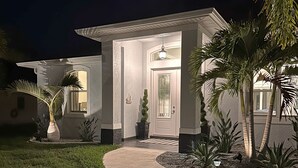 Une maison moderne dotée d’une entrée bien éclairée, d’une allée sinueuse et d’un jardin aménagé avec des palmiers et des arbustes.