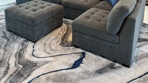 Un salon moderne avec un canapé d’angle et un pouf, une cuisine en arrière-plan et une grande fenêtre laissant entrer la lumière naturelle.