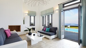 A modern living room with a sofa, a coffee table with a laptop, and a view of a pool through large windows. - Serenity View Villa with Caldera view, heated pool & hot tub in private garden (Thira - Santorini)