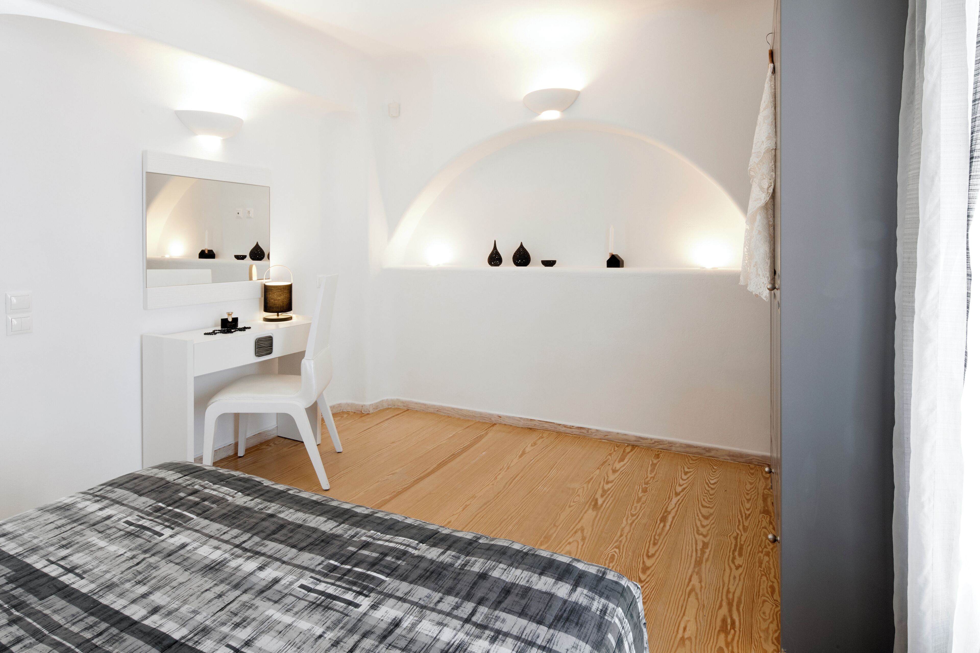 A modern bedroom with a bed, a desk, a chair, a mirror, and a wall-mounted shelf with decorative items.