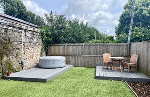A backyard with a stone wall, a wooden deck, a hot tub, and a table with chairs. - Detached Cottage with private garden and hot tub. (St Austell)