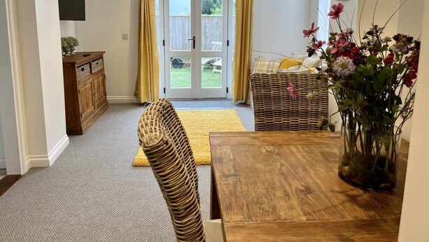 Een kamer met een houten eettafel, rieten stoelen, een rieten bank, een houten kast en een vaas met bloemen.