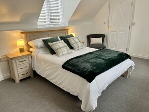 A bedroom with a bed, bedside tables, a chair, a window with blinds, and a door. - Detached Cottage with private garden and hot tub. (St Austell)