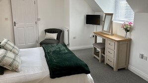 Een slaapkamer met een bed, een stoel, een bureau met spiegel, een televisie en een raam met zonwering.