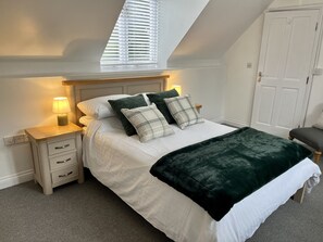 A bedroom with a bed, bedside tables, a lamp, a window with blinds, a door, and a chair. - Detached Cottage with private garden and hot tub. (St Austell)