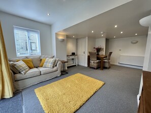 A modern living room with a sofa, a yellow rug, a dining area with wicker chairs, and a kitchen in the background. - Detached Cottage with private garden and hot tub. (St Austell)