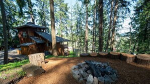 Mökki, jonka yhteydessä on nuotiopaikka, ja jonka ympärillä on tukkeja. Mökin sijainti on korkeiden puiden reunustamalla metsäisellä alueella.