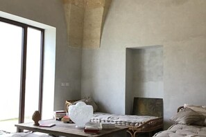Ein Zimmer mit großem Fenster, einem Holztisch mit Büchern und einem Dekorationsgegenstand, einem Bett mit gemustertem Überwurf und einem Gemälde an der Wand.