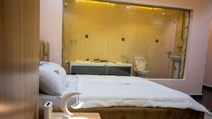 A bedroom with a large mirror, a bed with a white comforter, a bedside table with a lamp, and a view of a bathroom through the mirror. - Meraki Apartments (Akure)