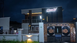 A modern building with a prominent 'Meraki' sign, featuring a large entrance with decorative metalwork and a gated driveway. - Meraki Apartments (Akure)