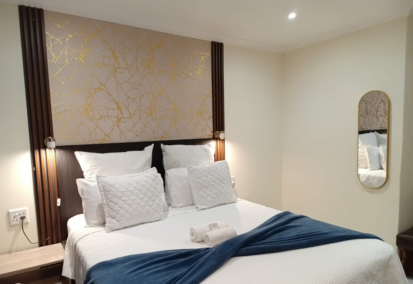 A neatly made bed with white bedding and a blue throw blanket, a wall-mounted mirror, and a decorative gold pattern on the headboard.
