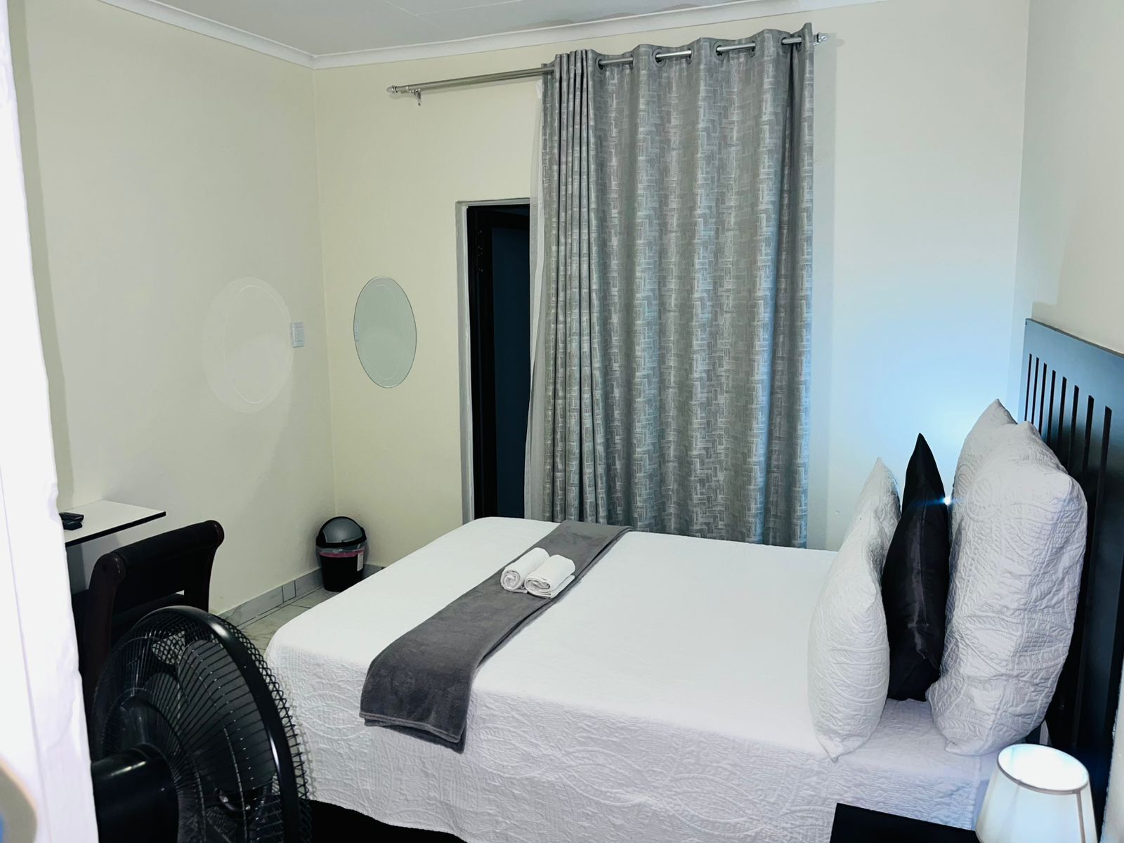 Quarto de hotel com cama, escrivaninha, cadeira, ventilador e janela com cortinas.