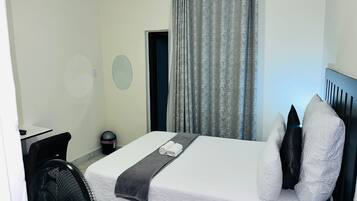 Quarto de hotel com cama, escrivaninha, cadeira, ventilador e janela com cortinas.