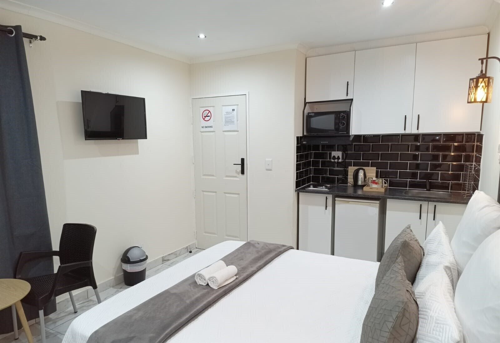 A compact hotel room with a bed, a small kitchenette, a TV, and a trash bin.