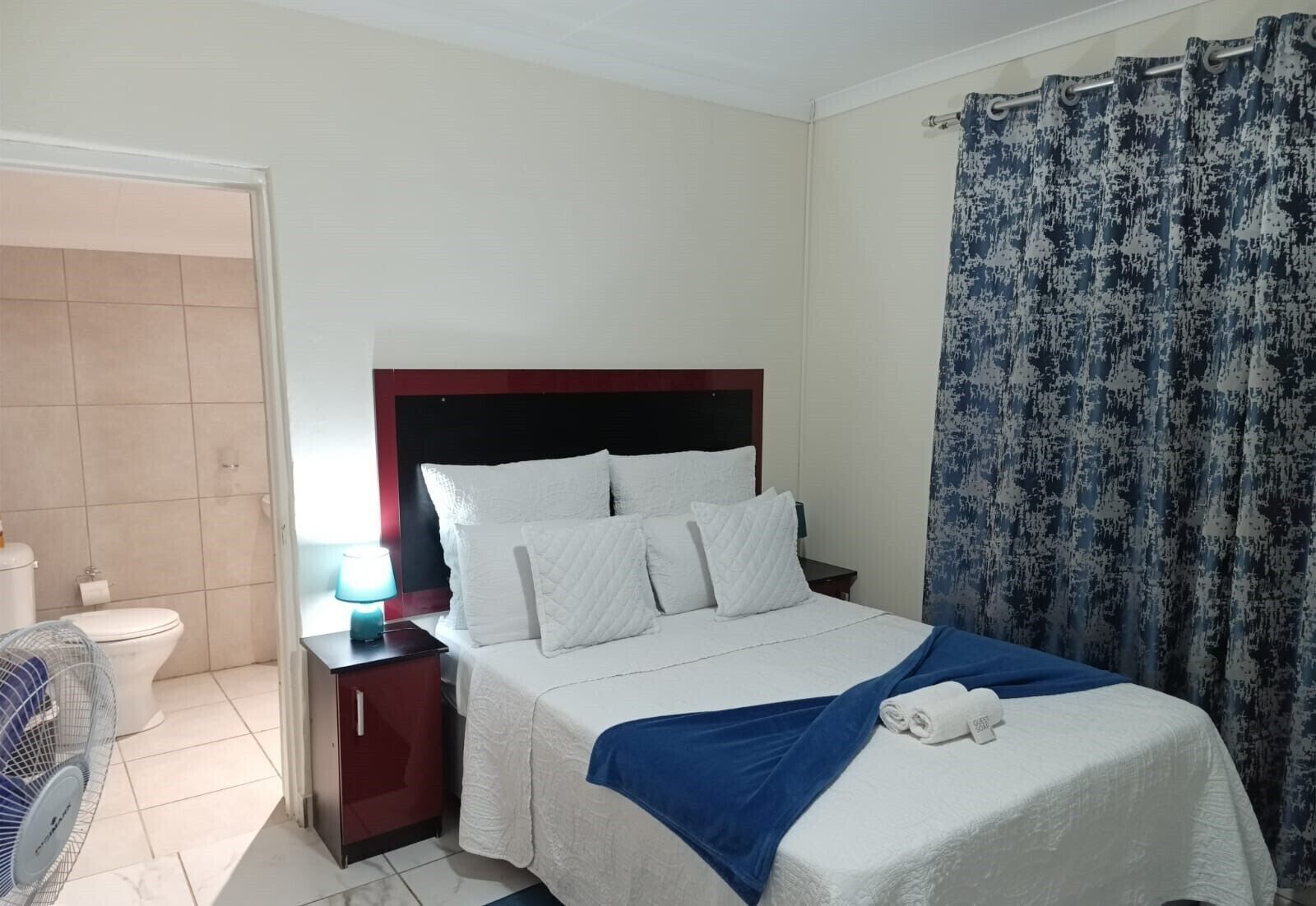 A hotel room with a bed, a nightstand, a lamp, a bathroom, and a curtain.