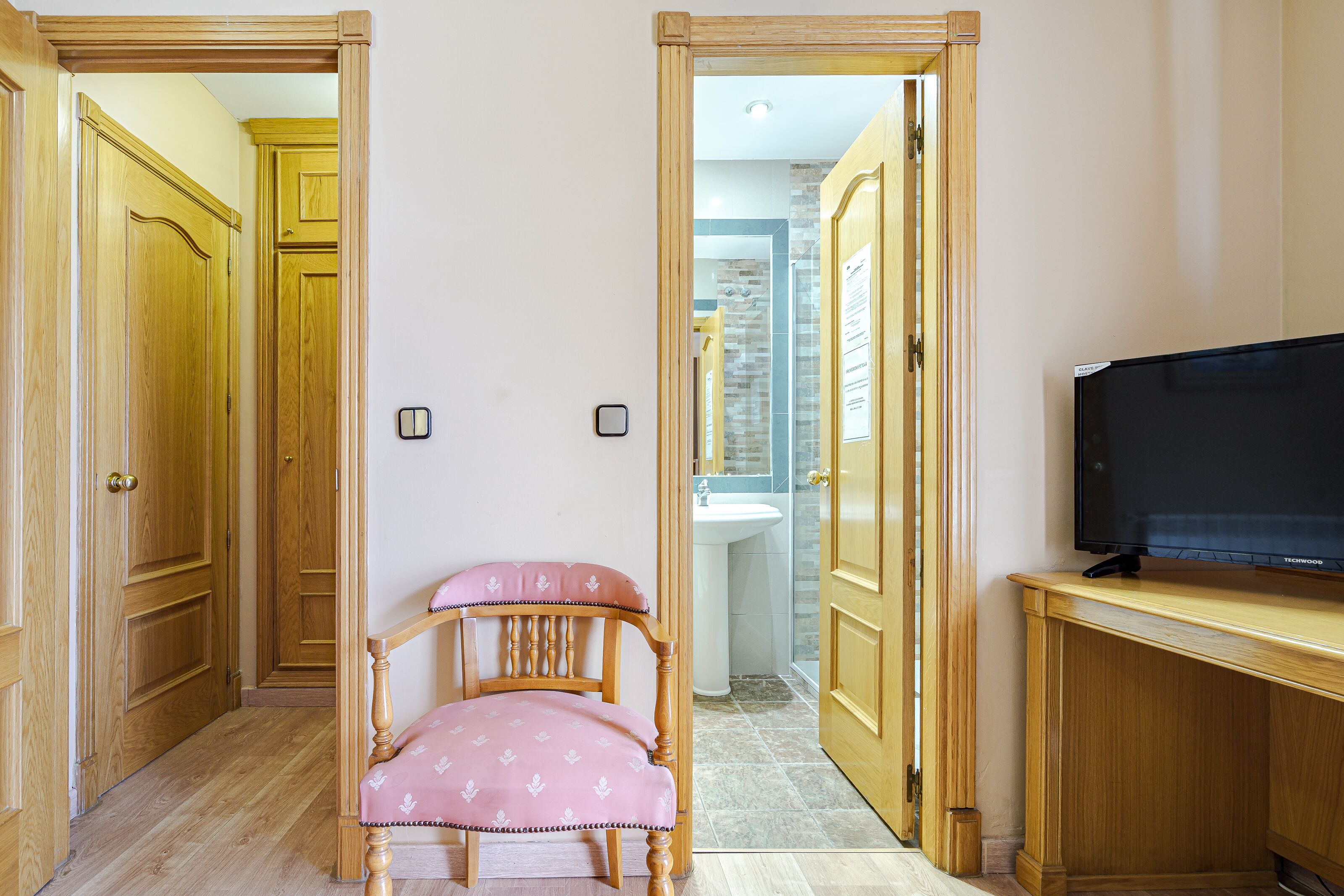 A room with wooden doors, a pink chair, a television on a wooden stand, and a bathroom visible through an open door.