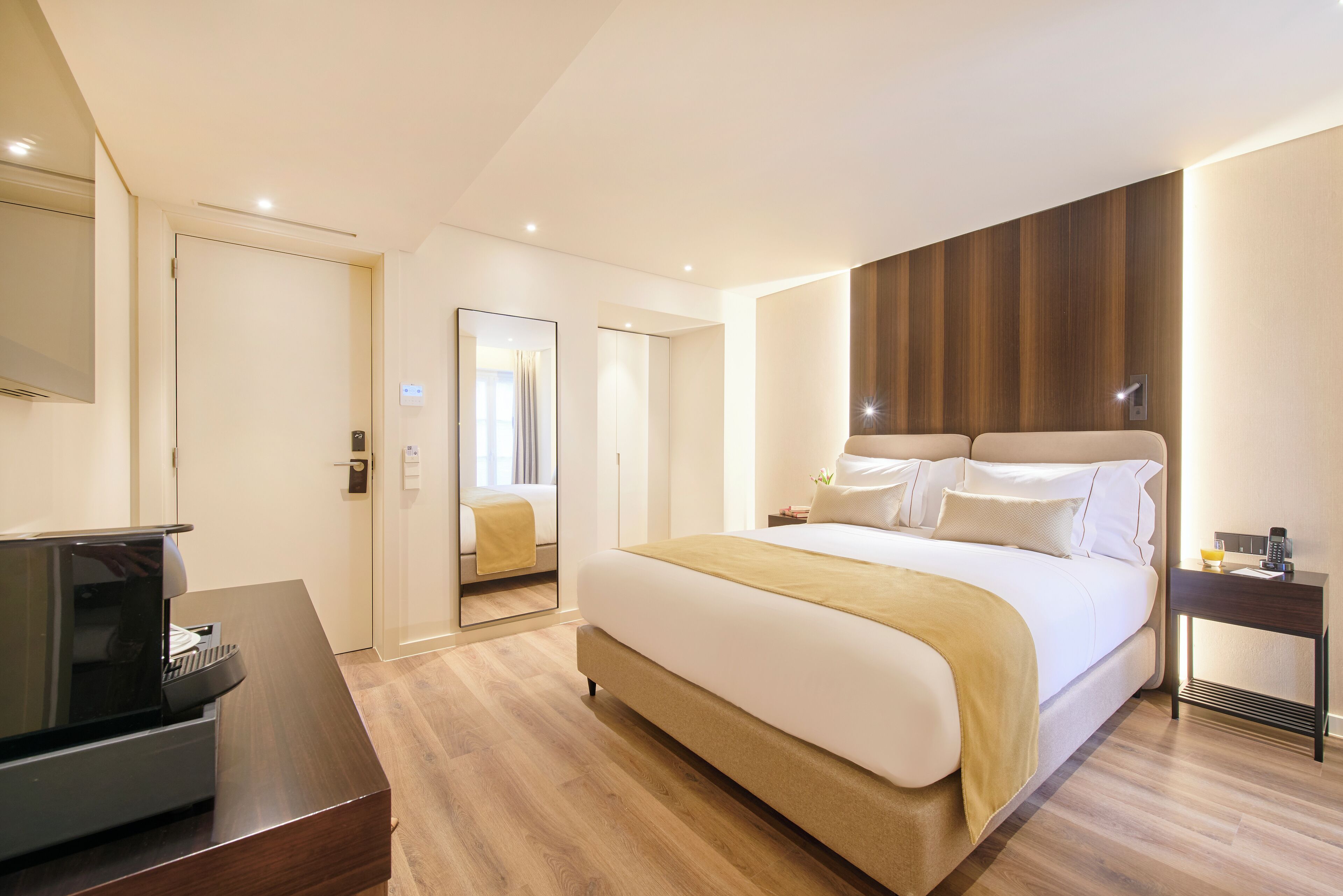 Una habitación de hotel moderna con una cama grande, un escritorio con una computadora portátil, una mesita de noche con una lámpara y un cabecero de madera.