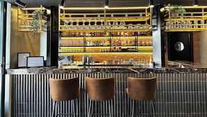 Zona de bar con tres taburetes, estantes con copas y botellas, y una pared de ladrillo.