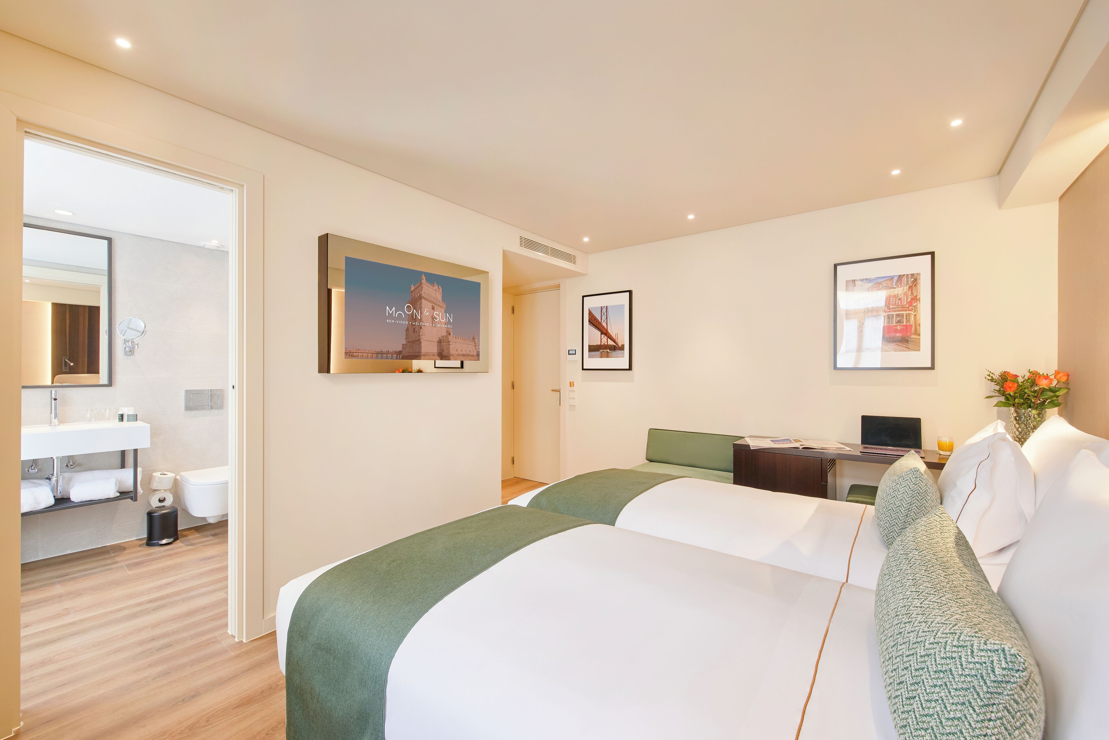 A hotel room with two beds, a desk, a chair, a small table, a mirror, a sink, and a toilet.