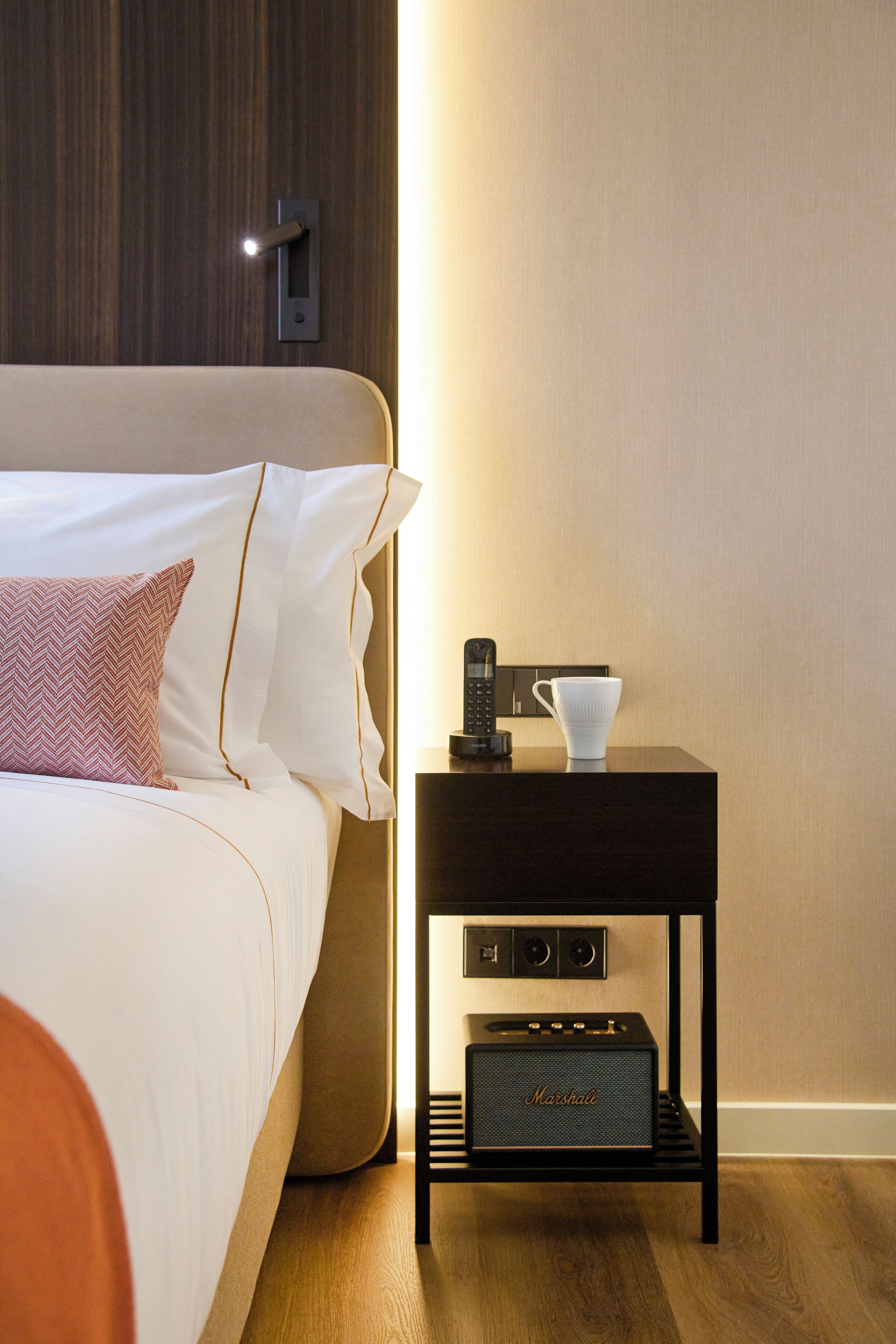 A neatly made bed with white linens and a pink patterned pillow, a bedside table with a black speaker, a white cup, and a black remote control.