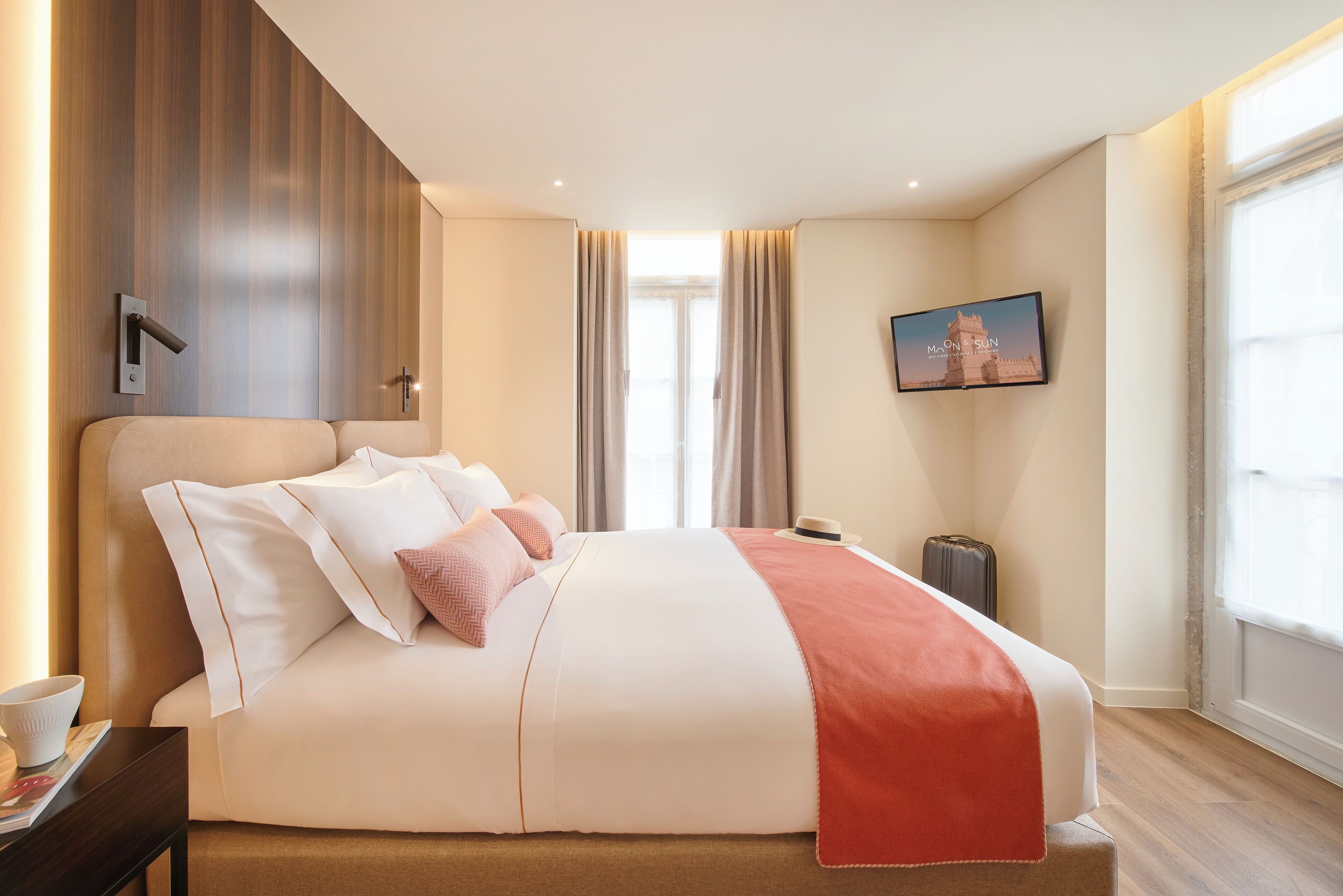 A hotel room with a large bed, a flat-screen TV mounted on the wall, a window with curtains, and a wall-mounted light fixture.