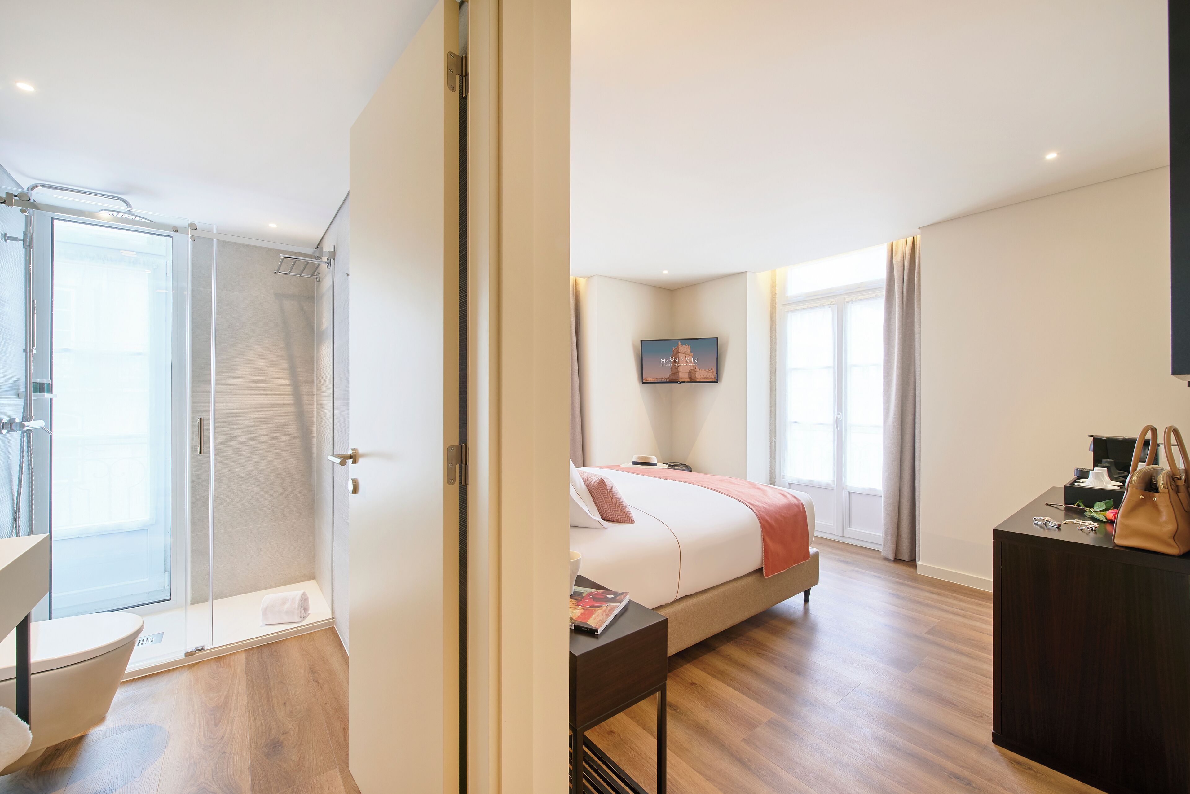 A modern hotel room with a glass shower enclosure, a bed with a red blanket, a flat-screen TV, and a wooden floor.