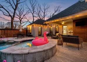 Un patio trasero con piscina, flamenco inflable, mobiliario exterior e iluminación con luces colgantes.