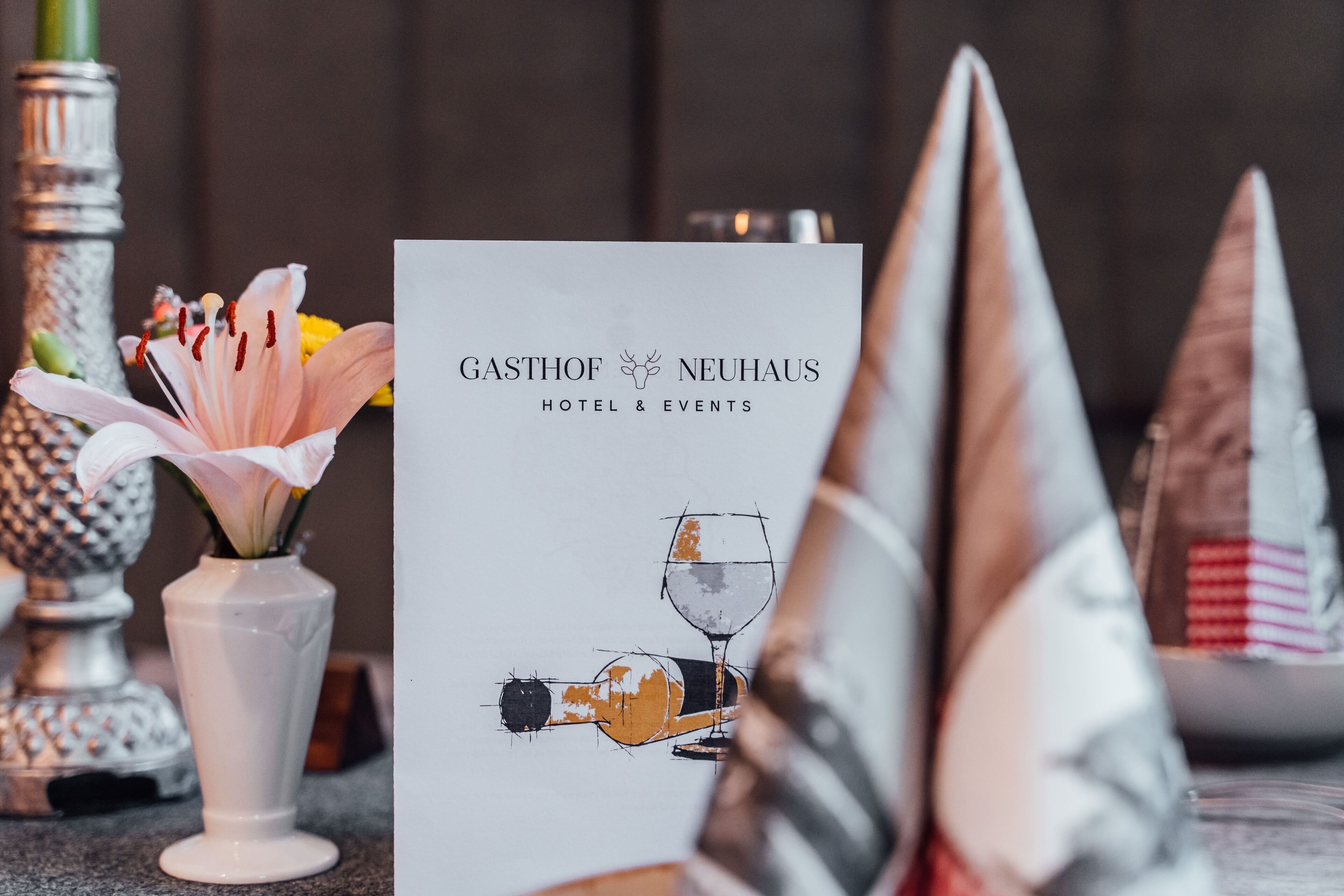 Une table est dressée avec un vase blanc rempli de fleurs roses, une bouteille de champagne et un verre à vin. Une pancarte indique : « GASTHOF NEUHAUS HOTEL & ÉVÉNEMENTS ».