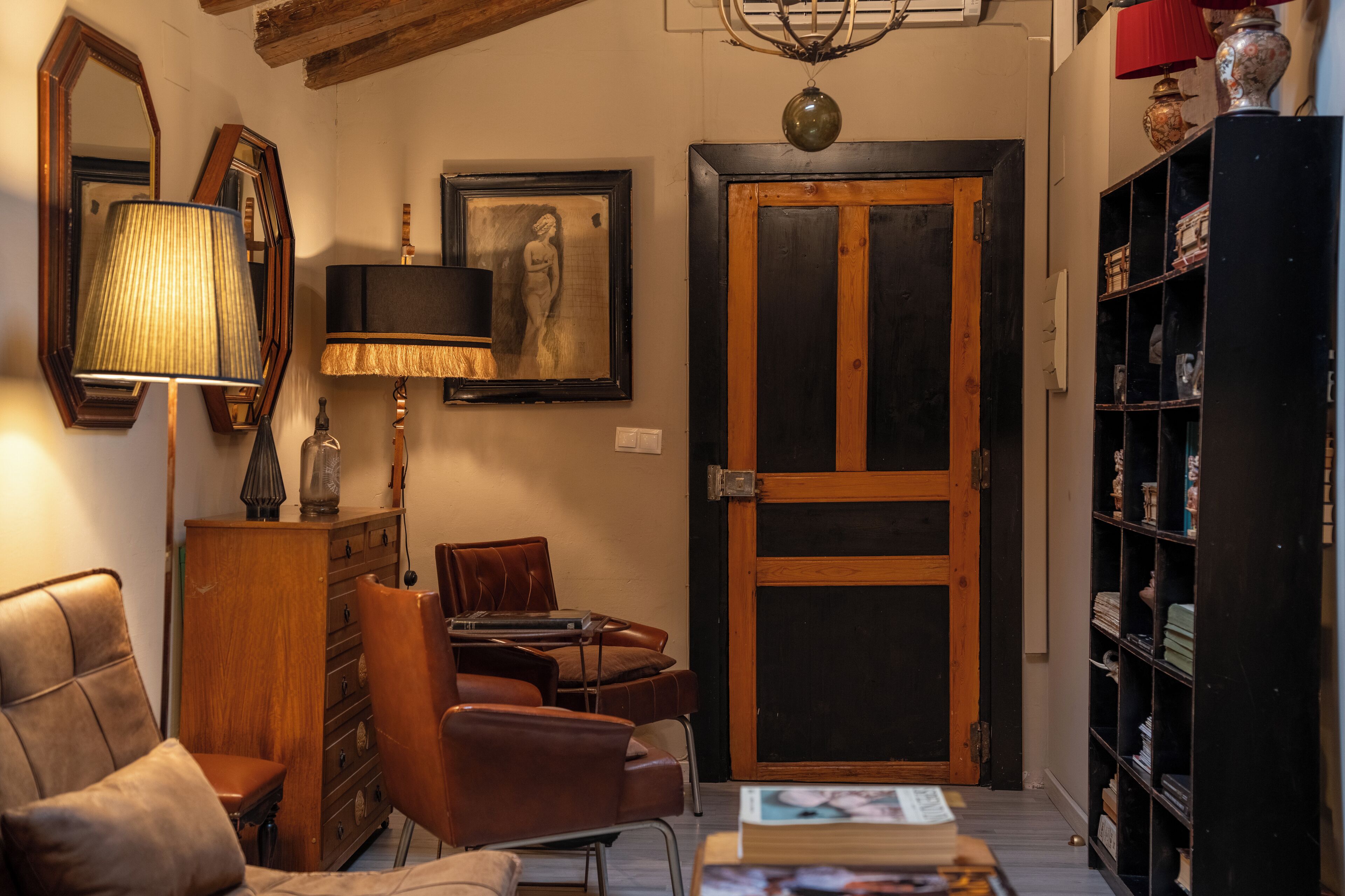 A cozy living room with a wooden cabinet, a leather armchair, a table with books, a lamp, and a door with a glass panel.