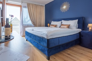A bedroom with a blue headboard, a wooden floor, a bedside table with a lamp, and a view of a house through a window. - Das Blaufränkisch Weinhotel & Restaurant (Deutschkreutz)