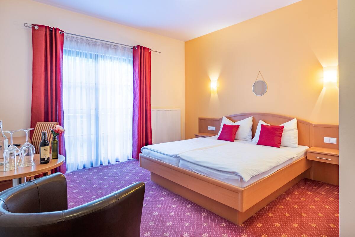 A hotel room with a bed, a desk with a chair, a table with wine glasses, and a window with curtains.