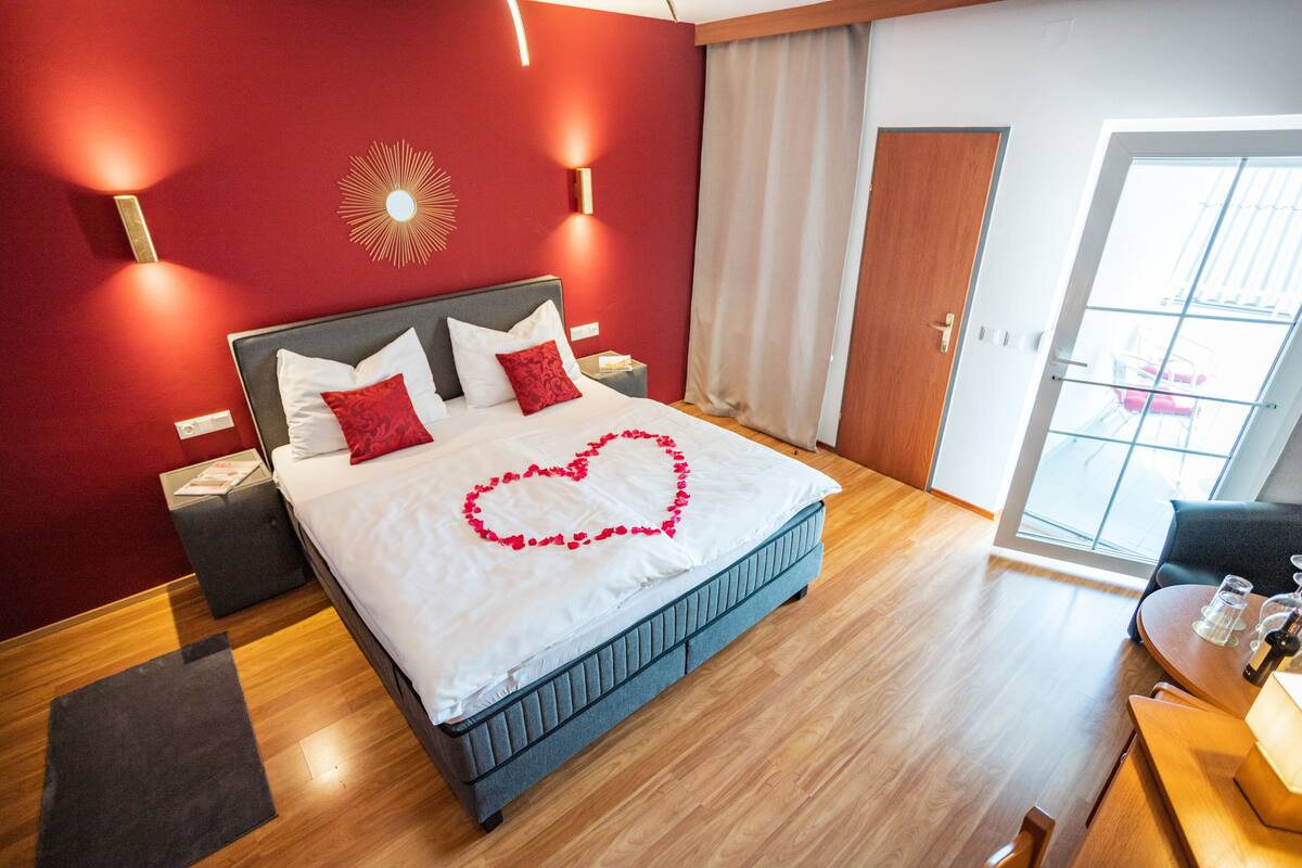 A modern hotel room with a large bed, a wooden floor, a red accent wall, and a view through a glass door.