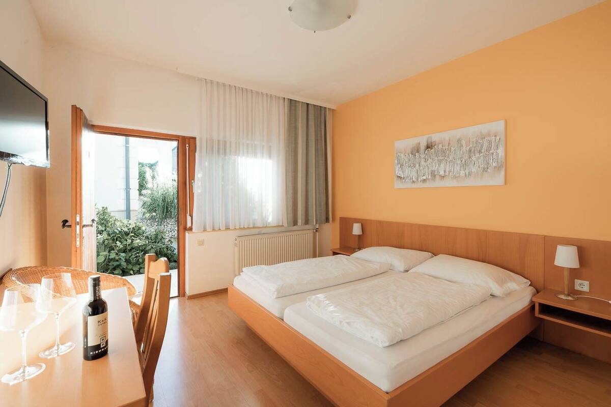 A hotel room with a bed, a table with wine glasses and a bottle, a window with curtains, and a painting on the wall.