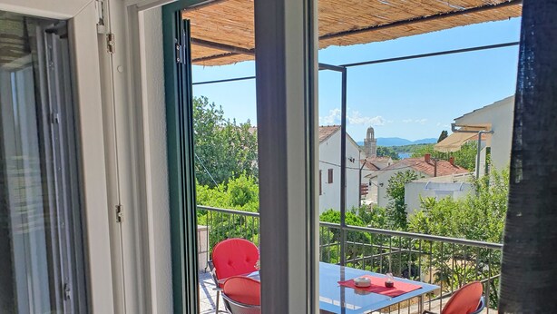 Ein Balkon mit Tisch und Stühlen, Blick auf Gebäude und Grünflächen, und ein Reetdach darüber.