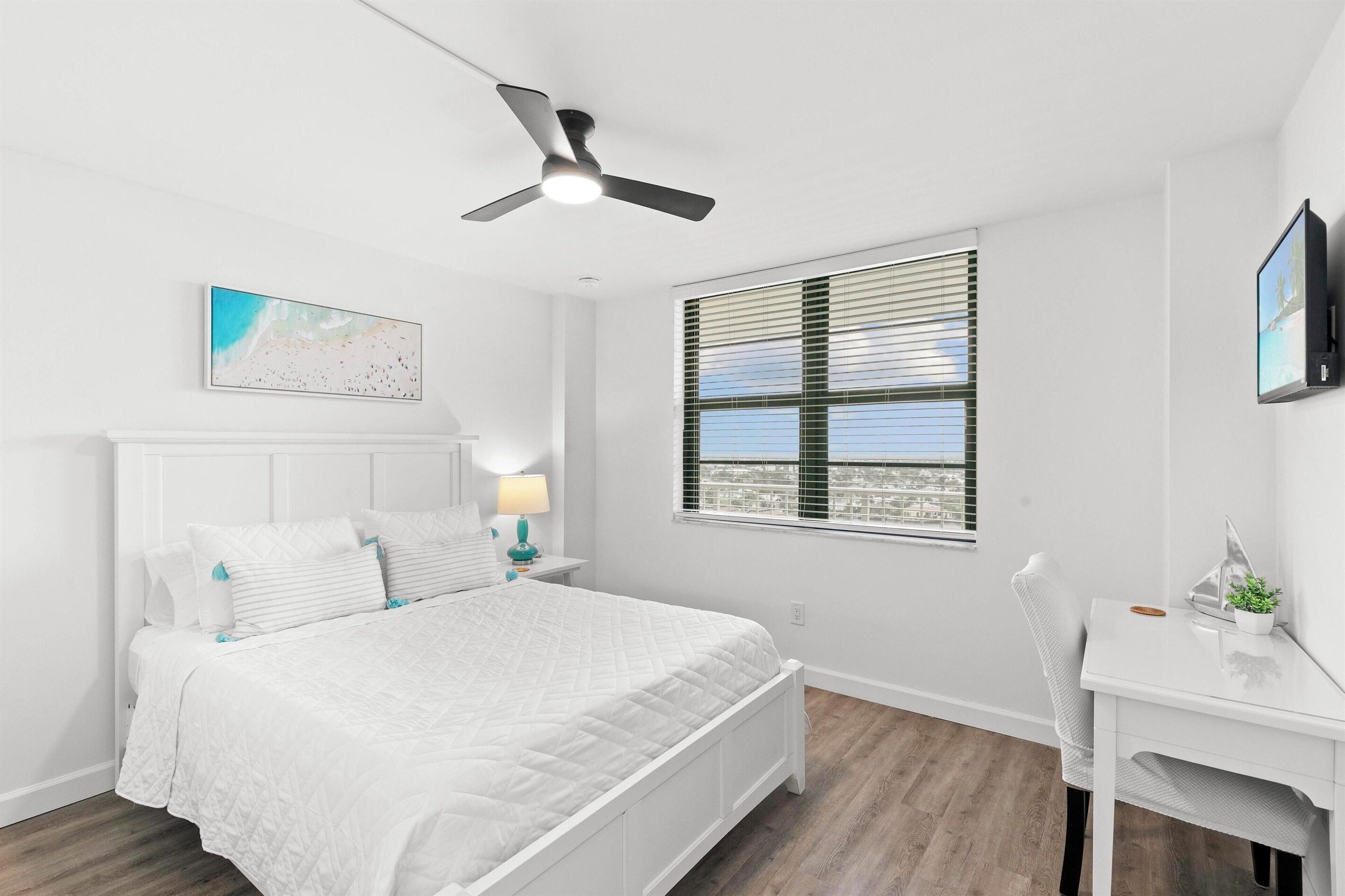 A bedroom with a bed, a desk, a ceiling fan, a window with blinds, and a wall-mounted TV.