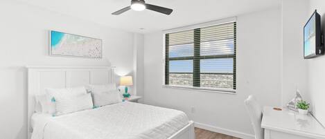 A bedroom with a bed, a desk, a ceiling fan, a window with blinds, and a wall-mounted TV.