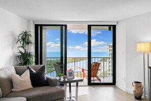 A modern living room with a sofa, a small table, a potted plant, and a view of the ocean through large sliding doors. - Award-Winning Oceanfront Condo, Sunset Views (Marco Island)